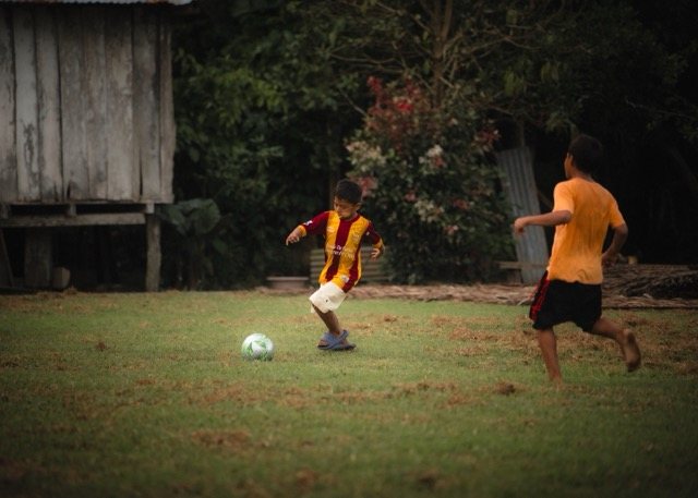 Two boys playing soccer on a grass field beside a wooden house
