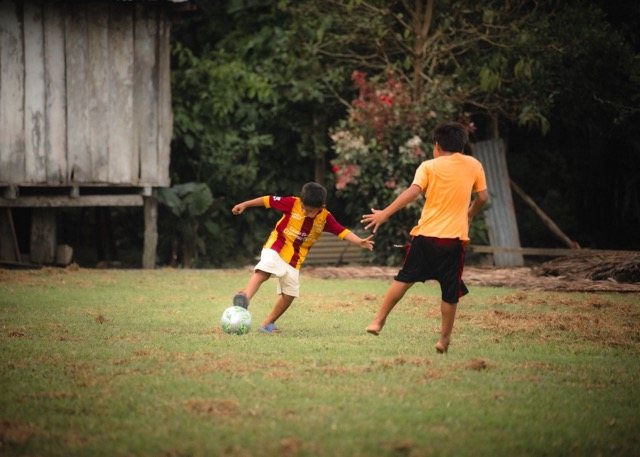 A boy in a red and yellow jersey striking the ball as a friend defends